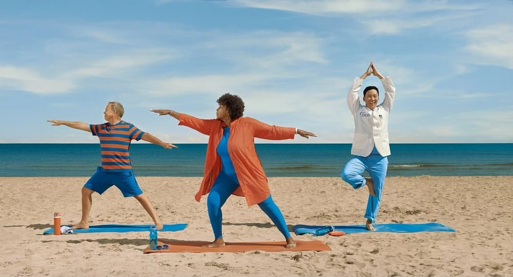 Three adults practicing yoga on a sandy beach, with the ocean and sky in the background, wearing fitness attire and a lab coat.